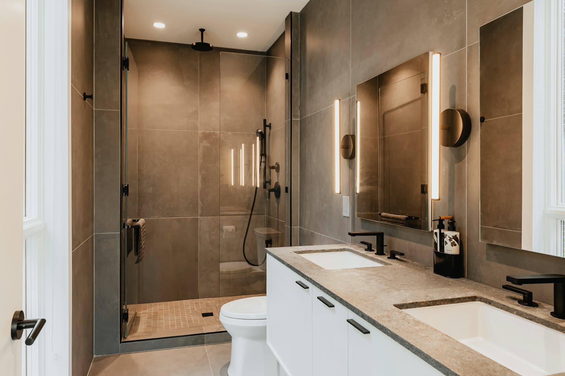 Yenilenmiş banyo; koyu tonlu büyük seramikler, cam panelli duş alanı ve beyaz lavabo dolabı